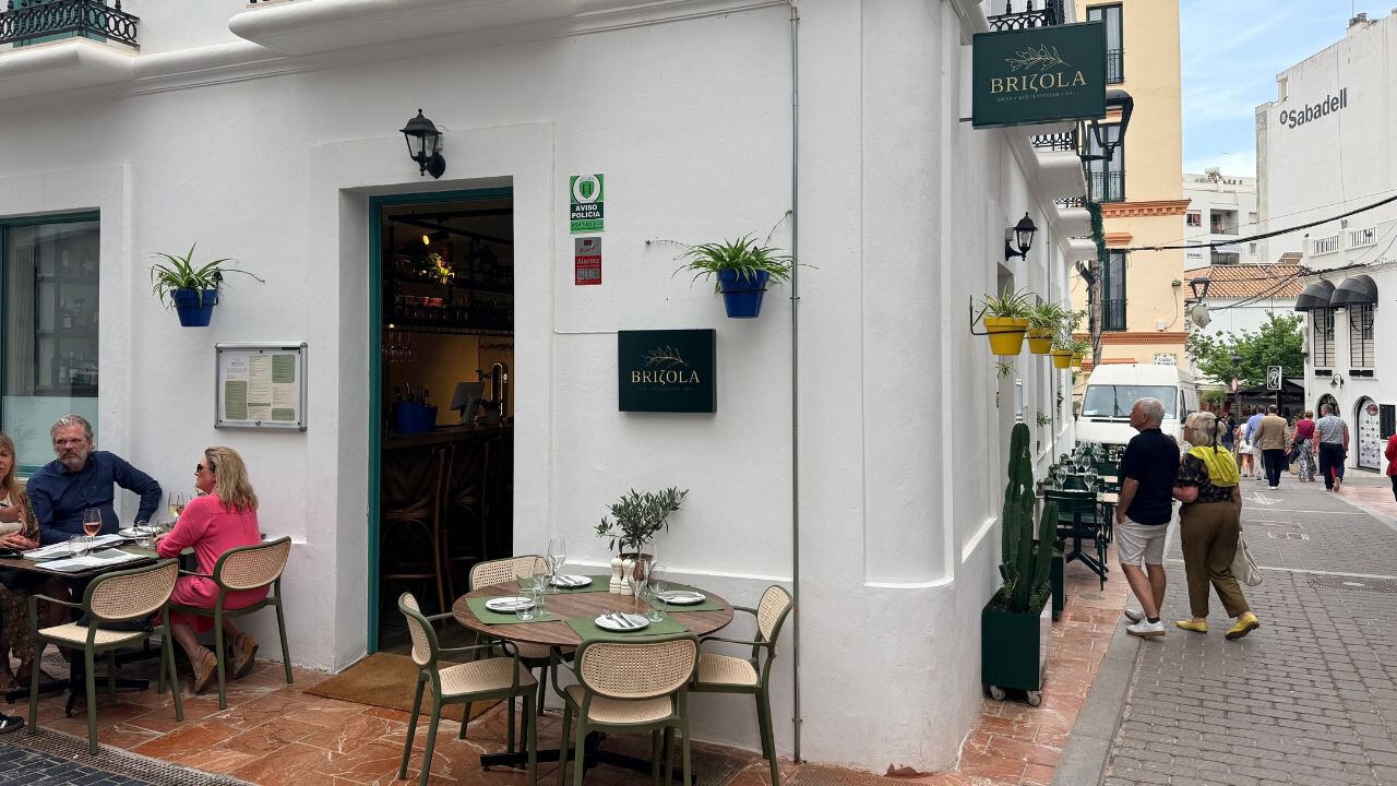 Brizola restaurant entrance with green signage, blue flowerpots and outdoor tables beside a pedestrian street in Estepona.
