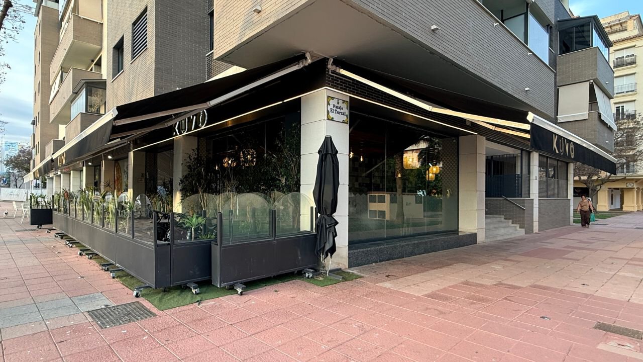 Kuvo restaurant on a street corner in Estepona, with black branded awnings and a plant-lined glass terrace.