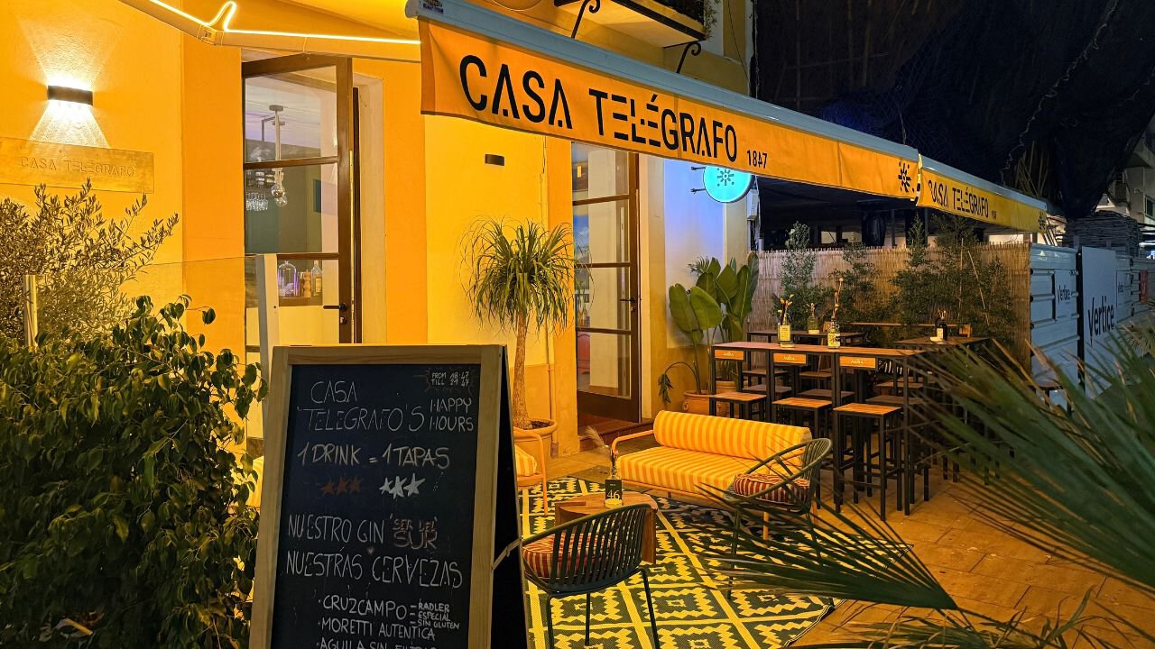 Illuminated Casa Telégrafo frontage in Estepona, with a yellow awning, outdoor seating, potted plants and a chalkboard.